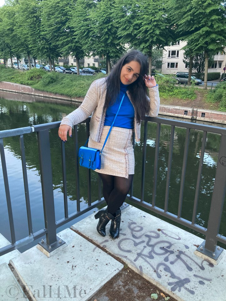 A beige skirt suit in French tweed worn with black accessories and an azure blue turtleneck and bag.
