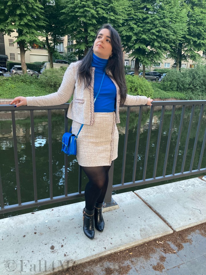 A beige skirt suit in French tweed worn with black accessories and an azure blue turtleneck and bag.