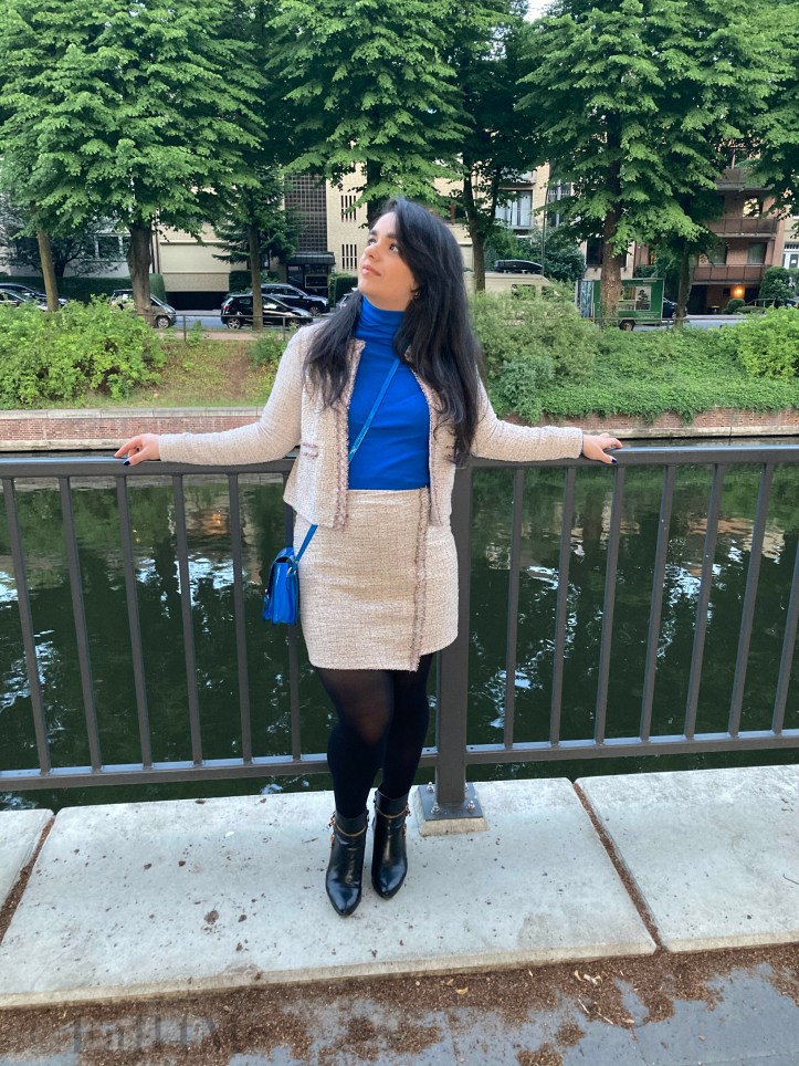 A beige skirt suit in French tweed worn with black accessories and an azure blue turtleneck and bag.
