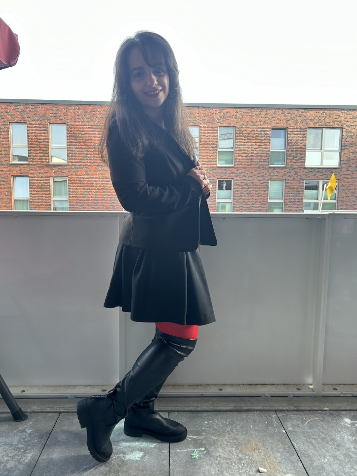 A pleated skirt making the skirt of the season worn with a black blazer, black over-knee-boots and red tights.